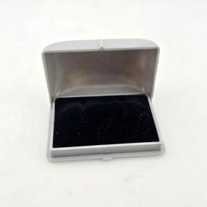 May include: A gray ink pad case, open to reveal a black ink pad. The case is rectangular with a hinged lid. The ink pad is a dark, velvety material. The case is sitting on a white surface.