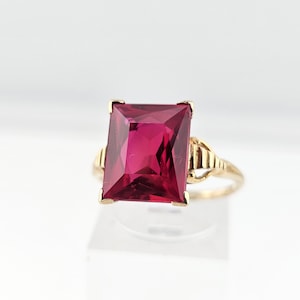 May include: A gold ring featuring a large, rectangular, deep pink gemstone. The ring has a classic design with a decorative band and a setting that securely holds the gemstone. The ring is set against a white background.