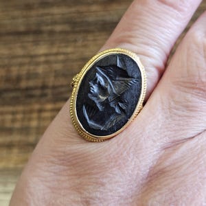 May include: A gold ring with an oval, black cameo depicting a profile of a person wearing a winged helmet. The ring has a detailed, textured gold band around the black cameo.