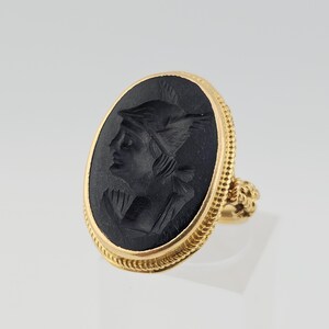 May include: A gold ring with an oval black cameo depicting a profile of a person wearing a winged helmet. The ring has a textured gold band and a decorative setting. The ring is a piece of jewellery.