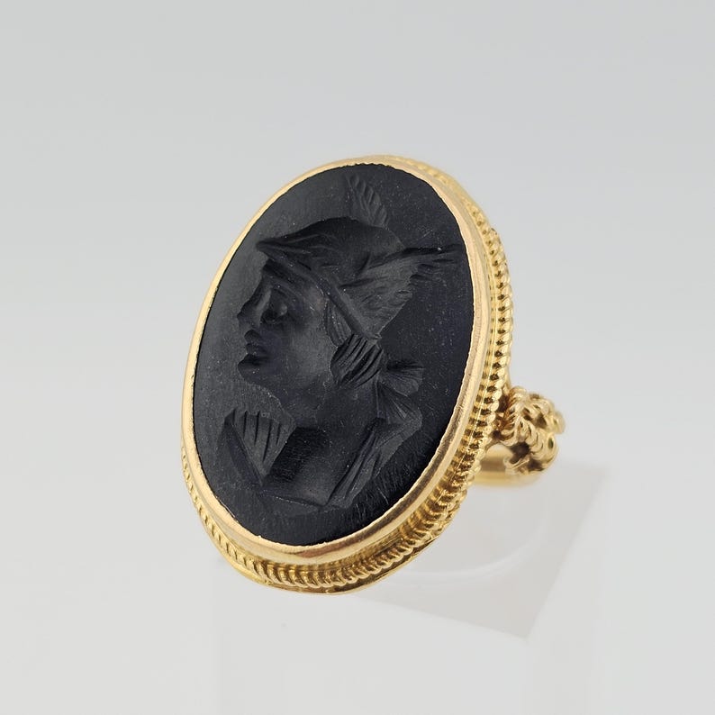 May include: A gold ring with an oval black cameo depicting a profile of a person wearing a winged helmet. The ring has a textured gold band and a decorative setting. The ring is a piece of jewellery.