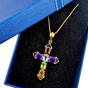 May include: A gold-colored cross pendant necklace with a variety of colorful gemstones. The gemstones are oval-shaped and include red, purple, blue, green, and orange hues. The necklace is presented in a blue gift box.