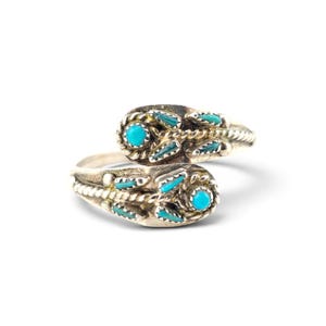 May include: A silver bypass ring featuring two rounded ends, each set with a central round turquoise stone surrounded by small, leaf-shaped turquoise inlays and twisted metal wire detailing along the band's surface.