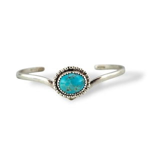 May include: A silver cuff bracelet featuring a vibrant oval turquoise stone set in a beaded bezel. The bracelet has a simple, open design with curved ends. The turquoise stone is a bright, eye-catching blue.
