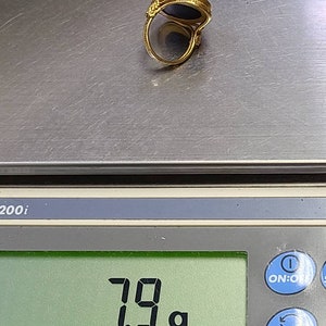 May include: A gold ring with a dark blue stone is displayed on a digital scale. The scale reads 7.9 grams. The ring has an ornate design around the stone and on the band.