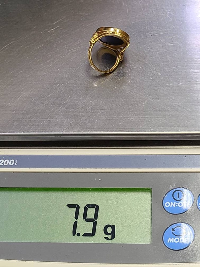 May include: A gold ring with a dark blue stone is displayed on a digital scale. The scale reads 7.9 grams. The ring has an ornate design around the stone and on the band.
