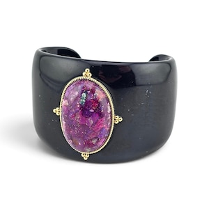 May include: A wide black cuff bracelet featuring a central oval-shaped stone with vibrant purple and magenta flecks. The stone is encased in a gold-toned metal setting with a twisted rope border and small decorative bead accents at the cardinal points.