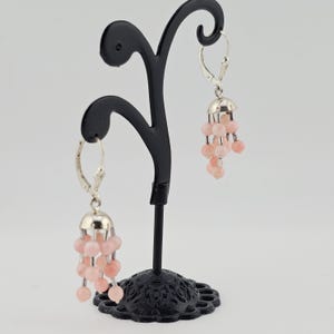 May include: A pair of dangle earrings with a silver-colored top and a cluster of light pink beads. The earrings are displayed on a black decorative stand against a white background. The earrings have a lever back closure.