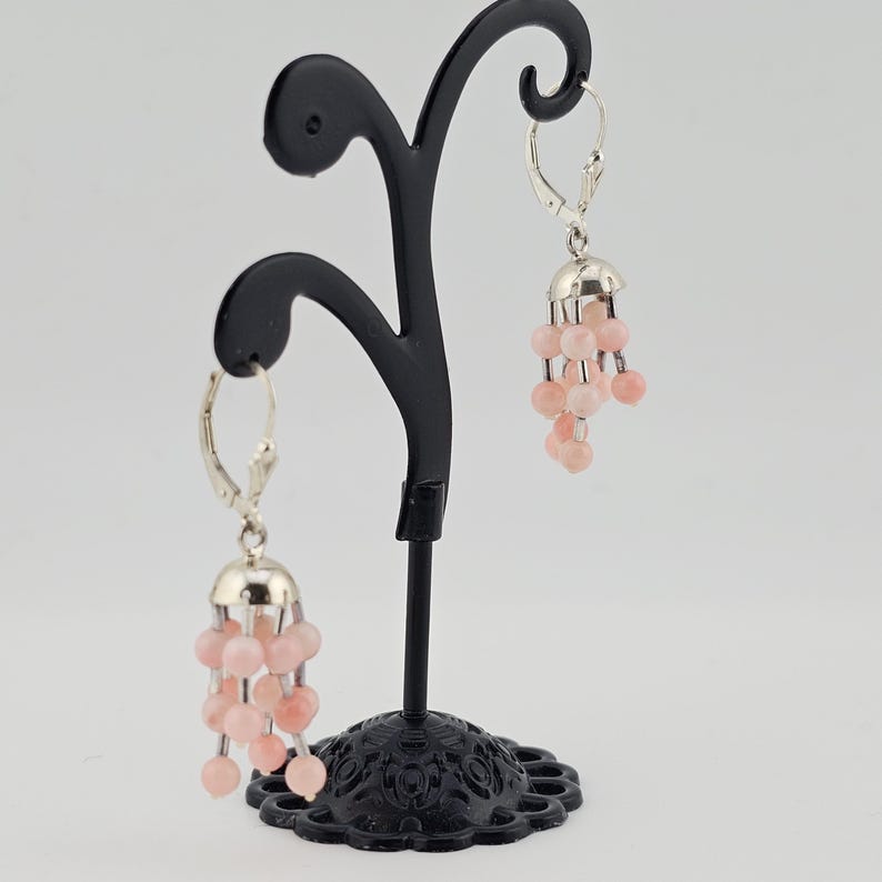 May include: A pair of dangle earrings with a silver-colored top and a cluster of light pink beads. The earrings are displayed on a black decorative stand against a white background. The earrings have a lever back closure.