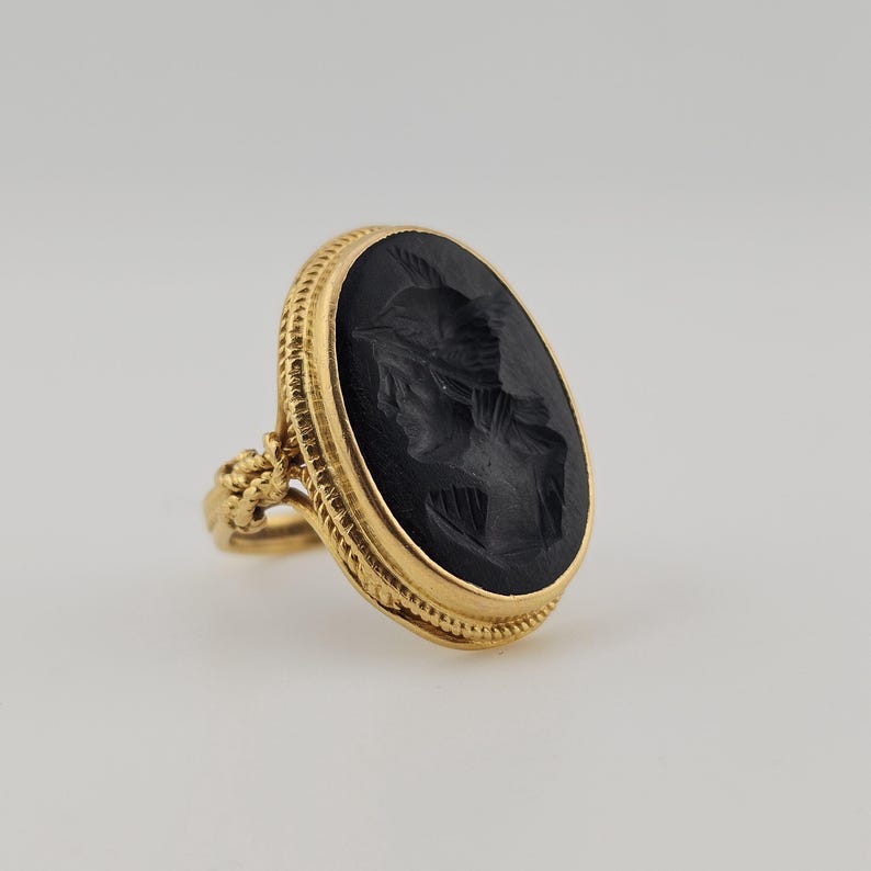 May include: A gold ring with an oval, black cameo depicting a carved profile. The ring has a detailed, textured band and setting. The cameo shows a classical figure. The ring is set against a neutral background.