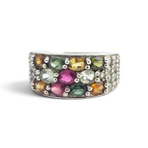 May include: A silver band ring featuring multiple rows of oval-cut gemstones in various colors including pink, green, yellow, and clear. The stones are set in a cluster arrangement across the front of the band, creating a multi-colored display.