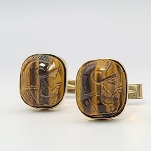 May include: A pair of gold cufflinks with a square tiger's eye stone set in each. The stones are carved with a detailed design.