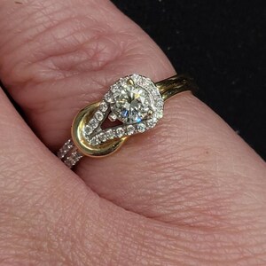 May include: A gold and diamond ring with a knot design. The ring features a large, round, clear diamond surrounded by smaller diamonds. The band is gold with additional small diamonds.