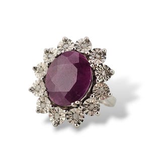 May include: A silver-toned ring featuring a large, oval-shaped, deep purple faceted gemstone at the centre. The stone is surrounded by a halo of twelve small, circular, textured silver-toned metal petals, each containing a tiny embedded clear stone.