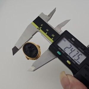 May include: A gold ring with a black oval stone, featuring a carved design. The ring is being measured with a digital caliper, showing a measurement of 2.475 cm. The ring has a detailed, textured band.