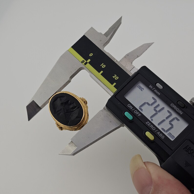May include: A gold ring with a black oval stone, featuring a carved design. The ring is being measured with a digital caliper, showing a measurement of 2.475 cm. The ring has a detailed, textured band.