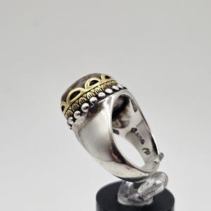 K&ouml;nnte beinhalten: Ein silberner Ring mit einem gro&szlig;en, ovalen Stein, der in einer goldenen Fassung sitzt. Der Stein ist von einem dekorativen Band aus goldenen und silbernen Perlen umgeben. Der Ring ist auf der Innenseite des Bandes mit "925" gekennzeichnet.