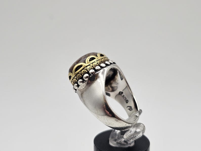K&ouml;nnte beinhalten: Ein silberner Ring mit einem gro&szlig;en, ovalen Stein, der in einer goldenen Fassung sitzt. Der Stein ist von einem dekorativen Band aus goldenen und silbernen Perlen umgeben. Der Ring ist auf der Innenseite des Bandes mit "925" gekennzeichnet.