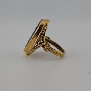 May include: A gold ring featuring an oval, dark-coloured stone. The ring has a detailed, rope-like design around the stone and on the band. The ring is set against a neutral background, highlighting its intricate details and craftsmanship.