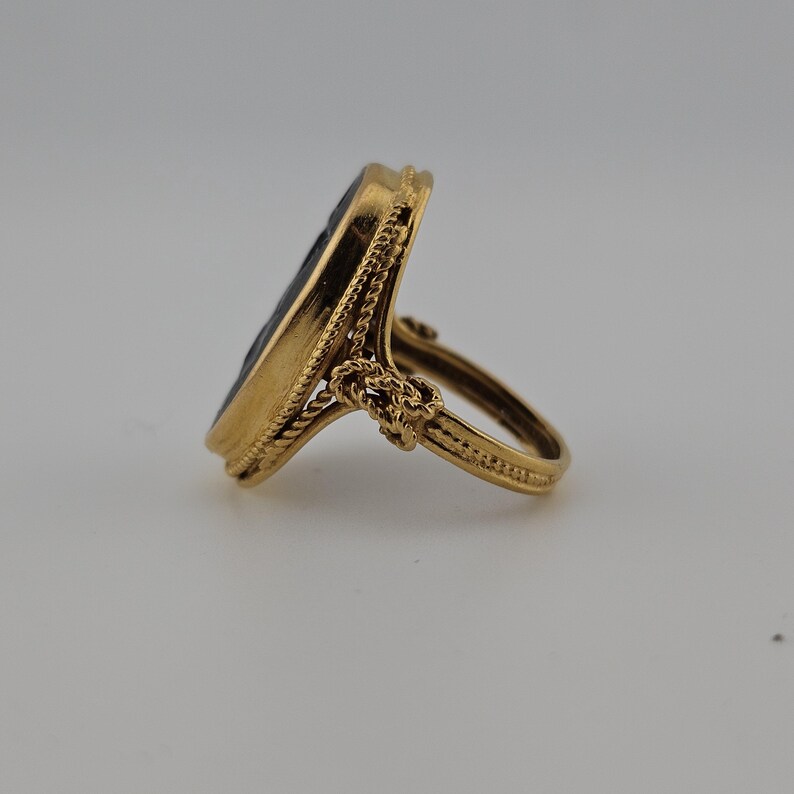 May include: A gold ring featuring an oval, dark-coloured stone. The ring has a detailed, rope-like design around the stone and on the band. The ring is set against a neutral background, highlighting its intricate details and craftsmanship.