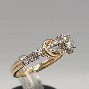 May include: A gold and silver engagement ring with a large, clear center stone. The ring features a knot design with smaller accent stones. The band is gold, and the setting around the main stone is silver.