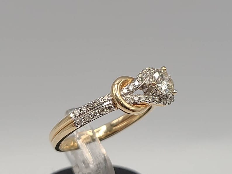 May include: A gold and silver engagement ring with a large, clear center stone. The ring features a knot design with smaller accent stones. The band is gold, and the setting around the main stone is silver.