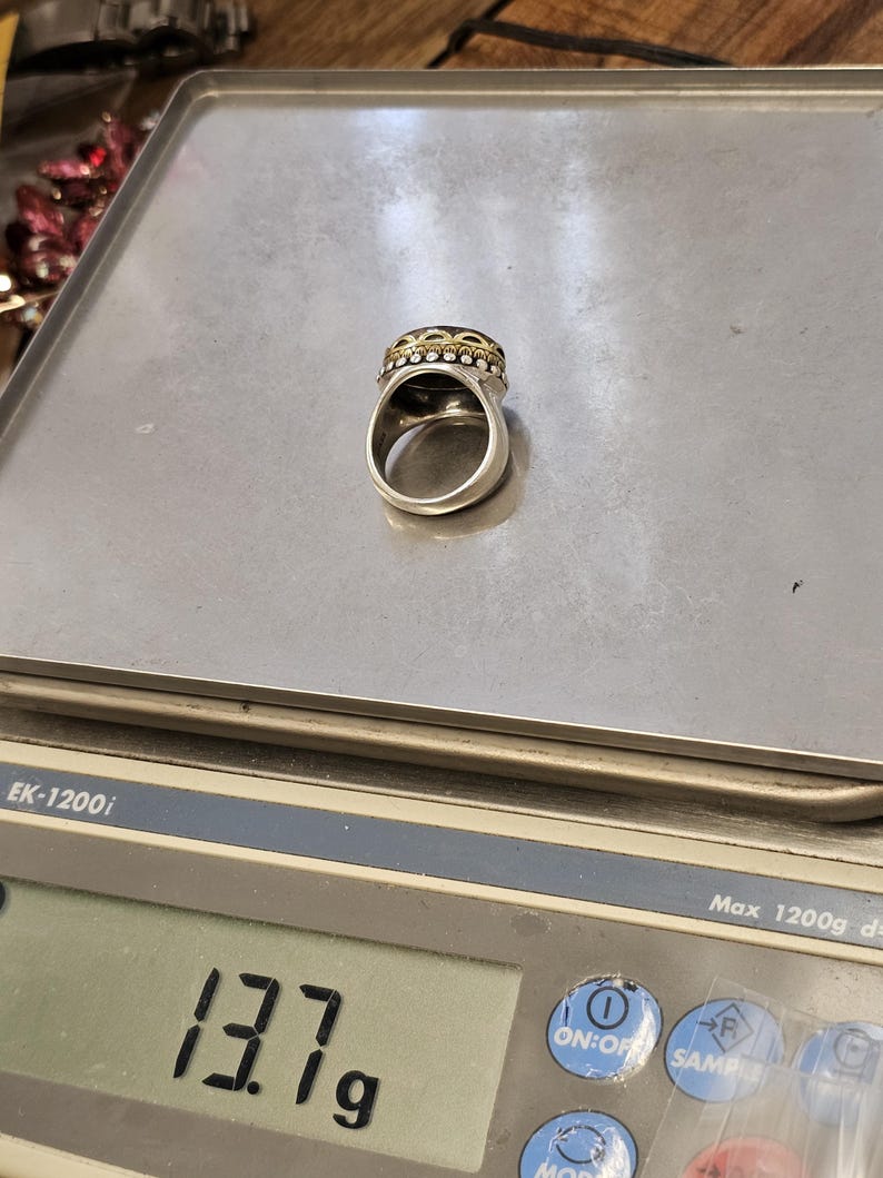 K&ouml;nnte beinhalten: Ein silberner Ring mit einem schwarzen Stein und goldenen Akzenten. Der Ring liegt auf einer digitalen Waage, die 13,7 g anzeigt.