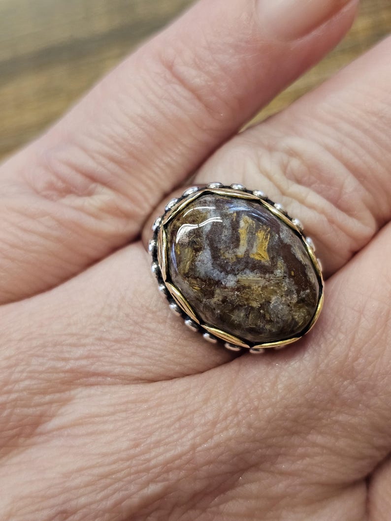 K&ouml;nnte beinhalten: Ein silberner Ring mit einem goldenen Band und einem braunen und wei&szlig;en ovalen Edelstein.