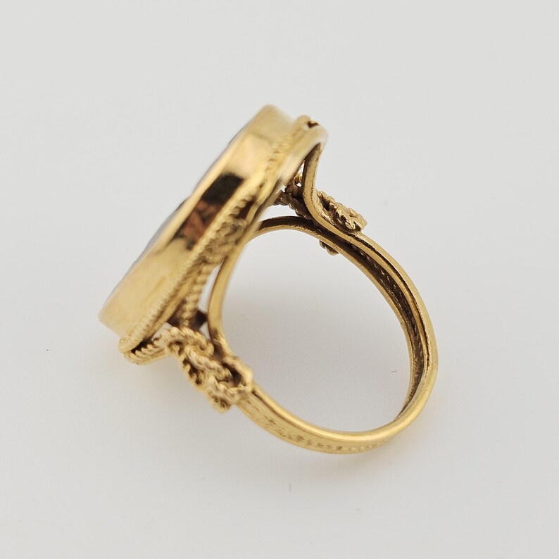 May include: A gold ring featuring an oval, dark-coloured stone. The ring has an ornate design with a textured band and decorative elements around the stone. The ring is set against a light background.