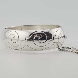 May include: A silver-colored bangle bracelet with an engraved swirl design. The bracelet has a hinged clasp with a safety chain. The design is repeated around the bracelet. The bracelet is a wide, oval shape.