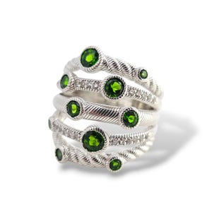 May include: A multi-strand silver ring featuring a twisted rope texture and multiple round, faceted green gemstones set along the bands. The design incorporates several smaller, clear, sparkling stones embedded between the green gems.