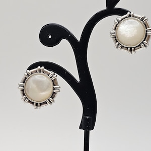 May include: A pair of silver earrings with white mother of pearl round insets. The earrings are on a black display stand.