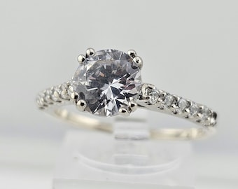 May include: A silver engagement ring with a large, round, clear gemstone as the centerpiece. The band is adorned with smaller, clear gemstones. The ring is set against a soft, neutral background, highlighting its sparkle and design.