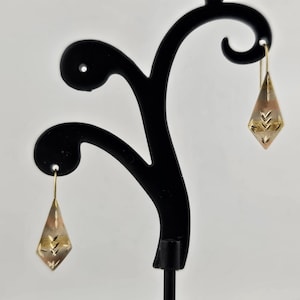 May include: A pair of diamond-shaped earrings with a gold-colored finish. The earrings have a geometric design with etched lines and are suspended from thin gold wires. The earrings are displayed on a black earring stand against a white background.