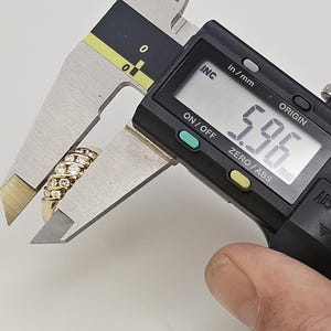 Puede incluir: Un anillo de oro con m&uacute;ltiples filas de peque&ntilde;as piedras preciosas transparentes se mide con un calibrador digital. El calibrador muestra una medida de 5,96 mm. El anillo est&aacute; sujeto por una mano.