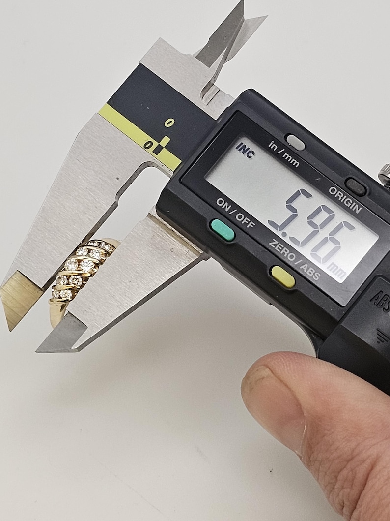 Puede incluir: Un anillo de oro con m&uacute;ltiples filas de peque&ntilde;as piedras preciosas transparentes se mide con un calibrador digital. El calibrador muestra una medida de 5,96 mm. El anillo est&aacute; sujeto por una mano.
