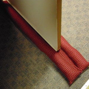 May include: A red doorstop with white polka dots is placed under a closed white door. The doorstop is made of fabric and is filled with padding.