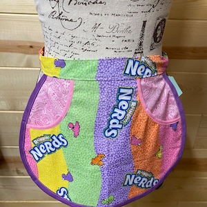 Può includere: Un grembiule rotondo e colorato con un motivo a strisce arcobaleno e le parole "Rainbow Nerds" stampate su di esso. Il grembiule è fatto di tessuto e ha un bordo viola.