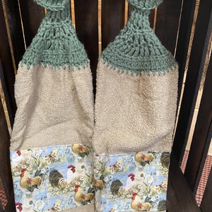 May include: Two hanging kitchen towels with a green crocheted top and a tan towel body. The towels feature a decorative band with a rooster and butterfly pattern. Each towel has a wooden button.