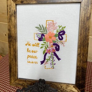 May include: Embroidered artwork in a wooden frame. The design features a cross adorned with colorful flowers, leaves, and purple ribbons. The text "He will be our peace. Micah 5:5" is written in gold. The frame has a dark brown finish.