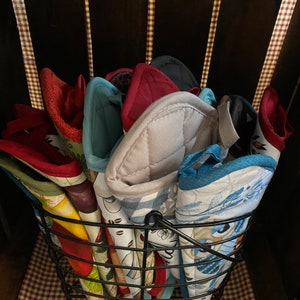 May include: A black wire basket filled with colorful quilted pot holders. The pot holders have various patterns and designs, including floral, geometric, and abstract.
