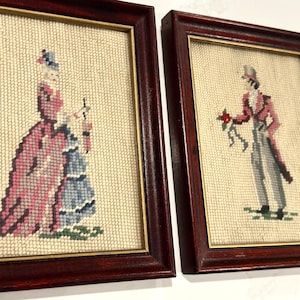 May include: Two framed needlepoint pictures. One depicts a woman in a pink dress and blue skirt, the other a man in a pink coat and gray pants. Both are framed in dark wood with gold trim.