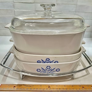 May include: A set of three white ceramic casserole dishes with blue floral designs. The dishes are stacked on a silver metal stand with three legs.