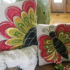 K&ouml;nnte beinhalten: Zwei dekorative Kissen mit handgemalten Designs. Ein Kissen zeigt eine gro&szlig;e rote, gelbe und gr&uuml;ne Blume mit einem schwarzen Zentrum. Das andere Kissen zeigt einen roten, gelben und gr&uuml;nen Schmetterling mit einem schwarzen K&ouml;rper.