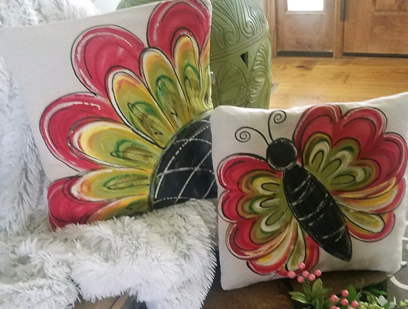 K&ouml;nnte beinhalten: Zwei dekorative Kissen mit handgemalten Designs. Ein Kissen zeigt eine gro&szlig;e rote, gelbe und gr&uuml;ne Blume mit einem schwarzen Zentrum. Das andere Kissen zeigt einen roten, gelben und gr&uuml;nen Schmetterling mit einem schwarzen K&ouml;rper.
