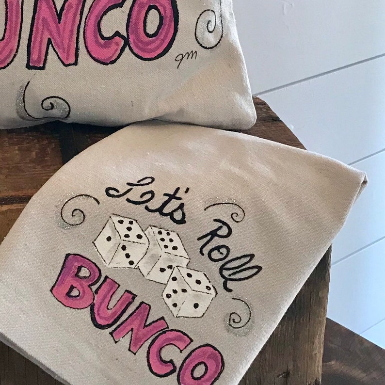 Peut inclure: Deux articles en tissu blanc cass&eacute; avec le mot "Bunco" en rose et en script noir. L'un des articles porte l'inscription "Let's Roll Bunco" avec des illustrations de d&eacute;s. Les articles sont sur une surface en bois.