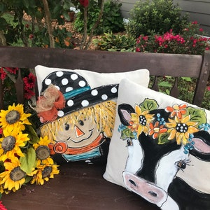 May include: Two decorative throw pillows with painted designs. One pillow features a scarecrow with a black and white polka dot hat and a straw brim. The other pillow features a black and white cow with a sunflower crown.