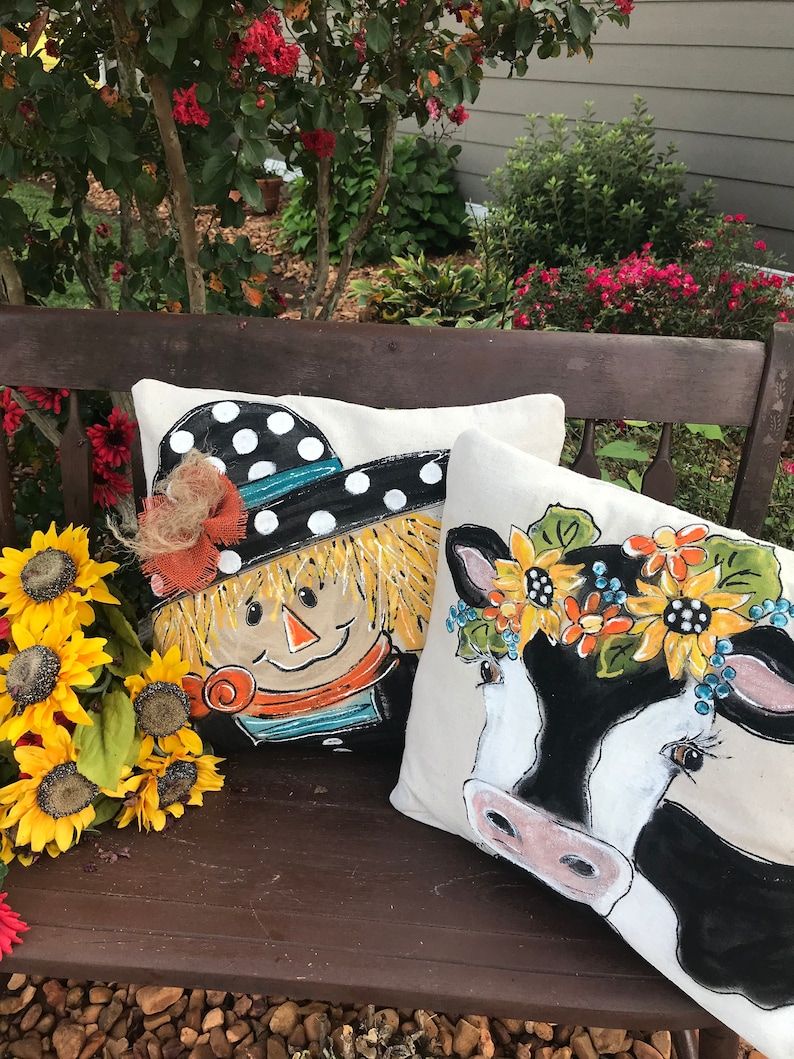 May include: Two decorative throw pillows with painted designs. One pillow features a scarecrow with a black and white polka dot hat and a straw brim. The other pillow features a black and white cow with a sunflower crown.