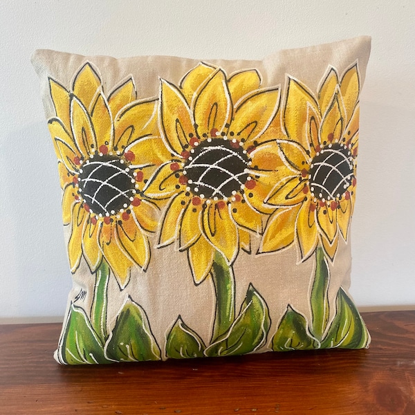 May include: A beige throw pillow with three yellow sunflowers painted on it. The sunflowers have brown centers and white accents.