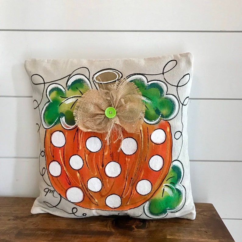 May include: A beige pillow with a hand-painted orange pumpkin with white polka dots. The pumpkin has a green stem and leaves and a burlap bow with a green button in the center.